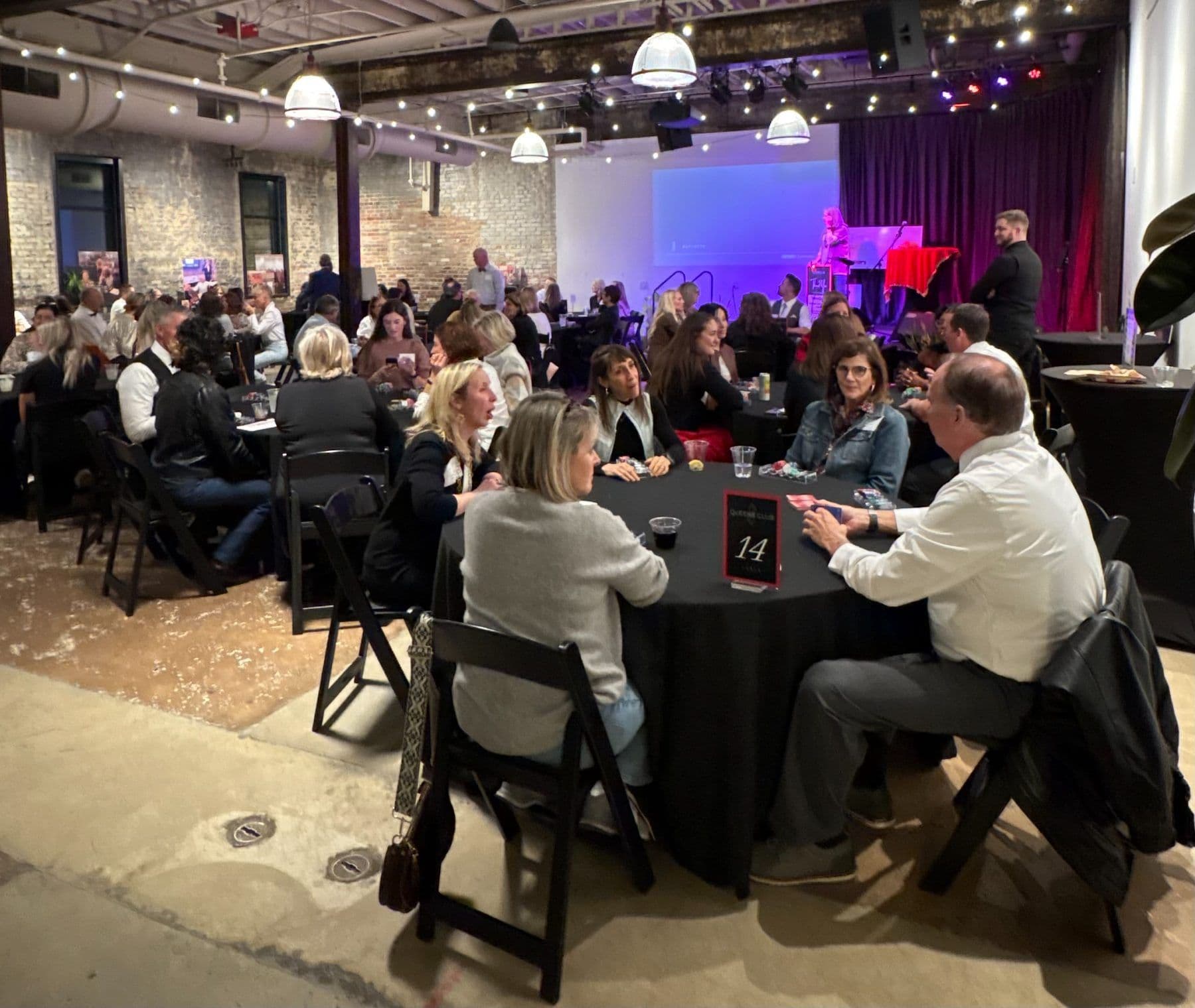 Guests seated at round poker tables in a brick-walled event hall under string lights