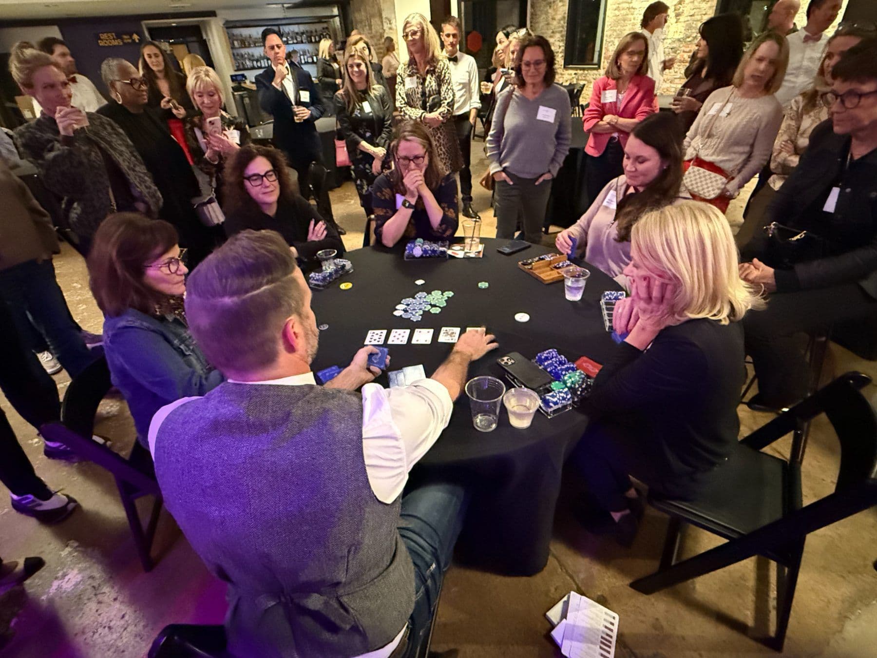 Women play a hand at a tournament poker table as a dealer lays out cards and a crowd looks on