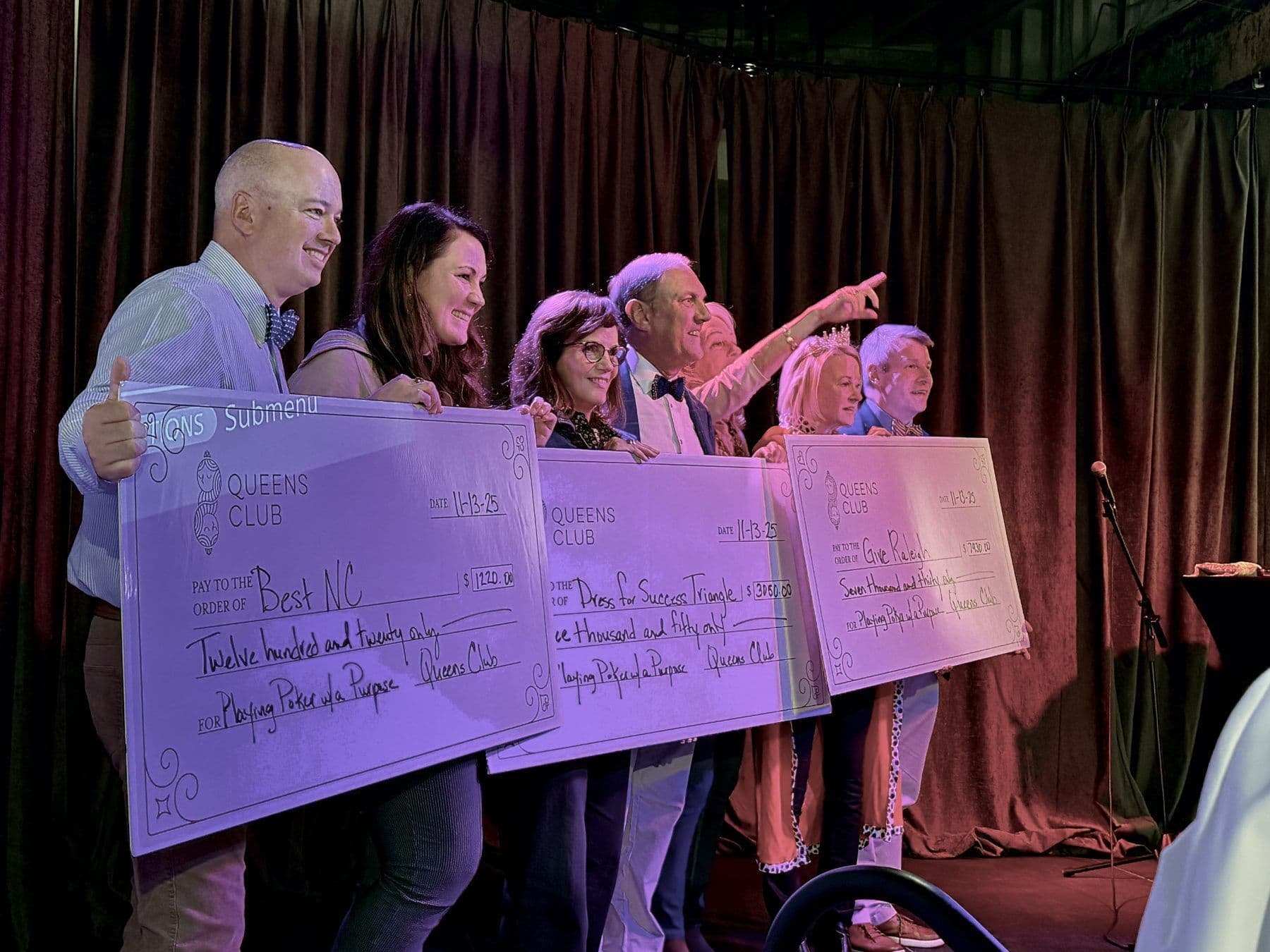 Final Table winners on stage holding oversized Queens Club checks made out to Best NC, Dress for Success Triangle and Give Raleigh
